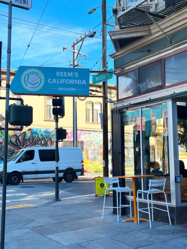 The exterior of Reem's, a Palestinian-owned restaurant in San Francisco. A white van passes on the road nearby, as diners can be seen inside a restaurant window, under a sign that reads: 