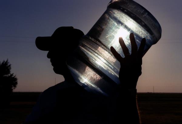 The silhouette of a man in a hat carrying a big bottle of water over his shoulder.