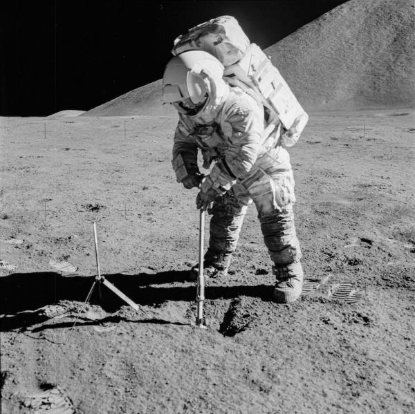 Astro<em></em>naut in bulky white spacesuit with large backpack holding a me<em></em>tallic instrument. He is standing in gray desolate terrain with hill behind him and black sky.