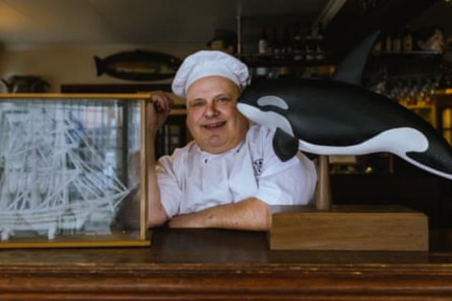 A heavily set man in a white chef’s hat poses in a restaurant next to a model of a ship and an orca 