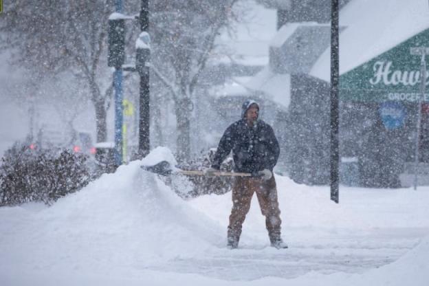 A man is seen holding a shovel amid a snowy street, next to a small snowbank.