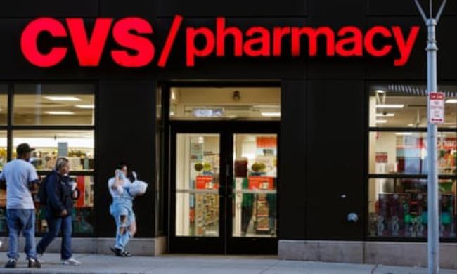The outside of an urban CVS pharmacy, with red letters lit up and a black building facade, with three people walking toward the front doors.