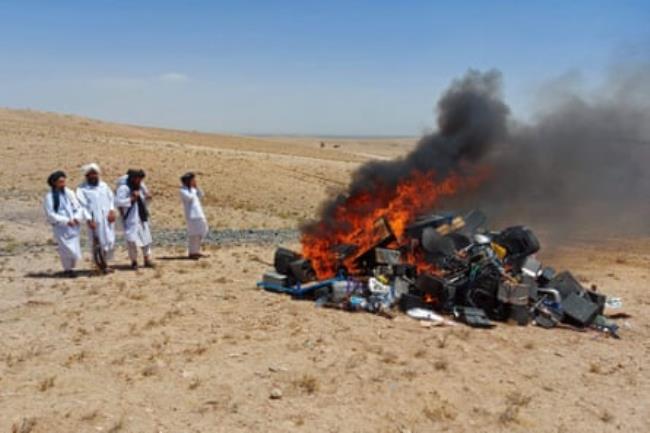 Four Taliban watch a pile of musical instruments and equipment burn in a large fire.