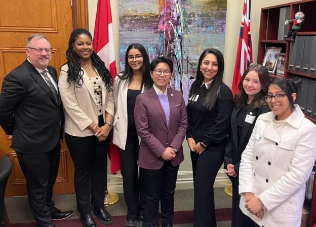A group of politicians pose with post-seco<em></em>ndary students indoors, with the Canadian and o<em></em>ntario flags along with an abstract painting behind them. 