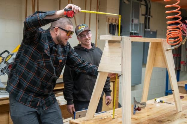 Two people measure a wooden bench in a woodworking shop.