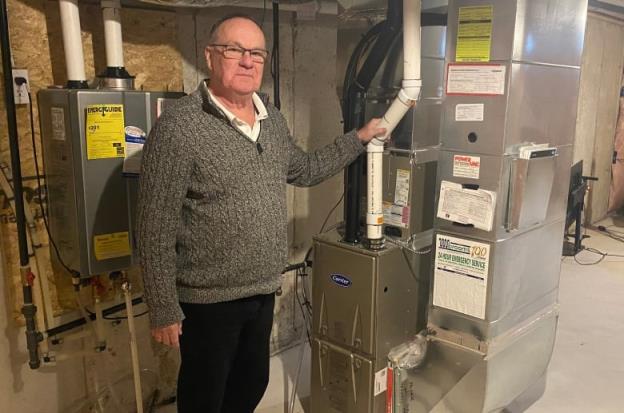 An older man with glasses wearing a grey sweater and dark pants stands in a ba<em></em>sement with his hand on a furnace pipe.