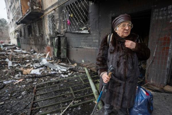 An elderly woman standing in front her damaged house after Russia launched 10 missiles on Kyiv. The wall is blackened and there's debris on the ground behind her. She's holding a broom.