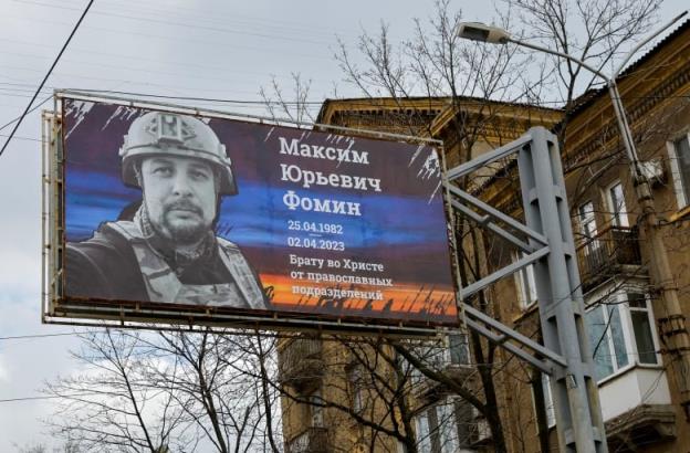 A billboard near a low-rise building is shown, displaying the picture of a bearded man wearing a helmet and a military vest.