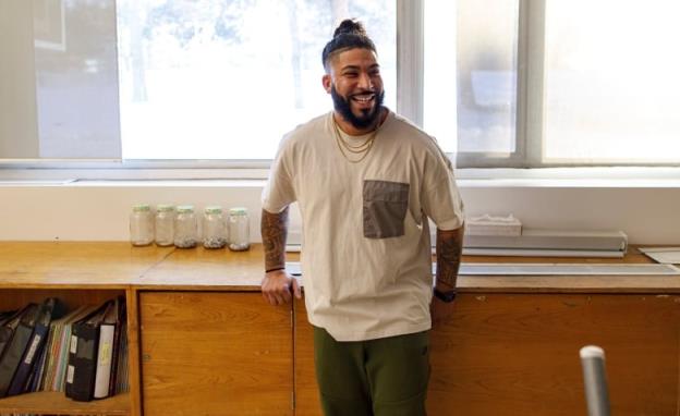 A male teacher looks and laughs at someone off-camera to the right, while leaning back against wooden cupboards in his classroom.