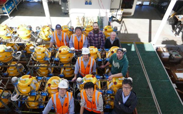 A group of nine people wearing hard hats and hi-vis vests gather next to large yellow cylindrical devices in me<em></em>tal f<em></em>rames on the deck of a ship.