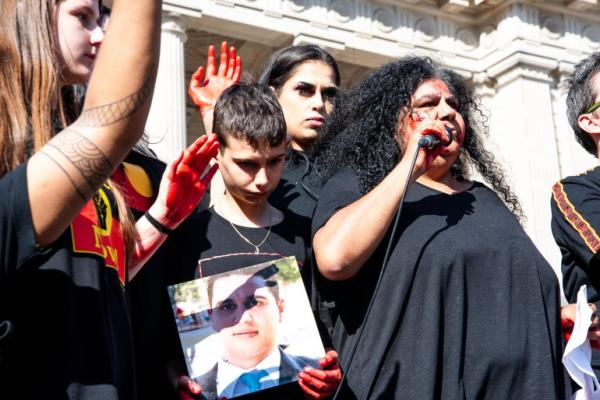 Do<em></em>nnis Kerr. She is at a rally and speaking into a microphone. She has far curly hair and is wearing a black shirt.