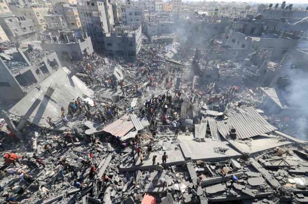 An overhead shot showing a large crowd of people standing in the middle of a large pile of debris in an urban neighborhood in the Gaza Strip.