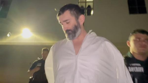 Stephan Sterns, a white man with dark hair and a graying beard, with his hands behind his back as he is escorted by a uniformed sheriff.