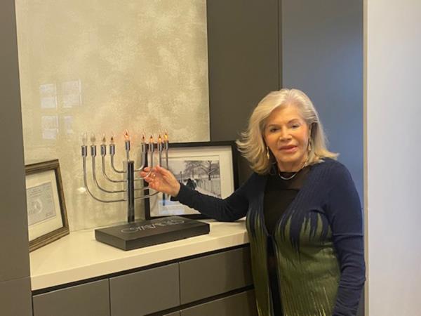 Lucy Lippiner resting her hand on a menorah sitting on a book case, next to f<em></em>ramed photos