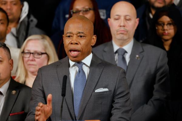 Mayor Eric Adams standing at press co<em></em>nference speaking with micropho<em></em>nes and NYC officials around him including a female police officer in uniform.