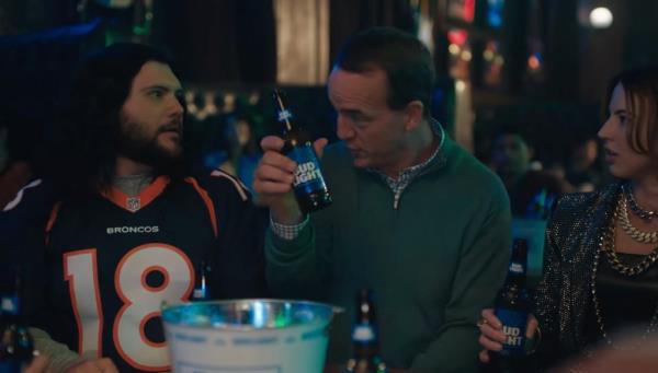 A man holding a bottle of Bud Light beer with Peyton Manning and other celebrities in the background at a party during the Super Bowl advertisement.