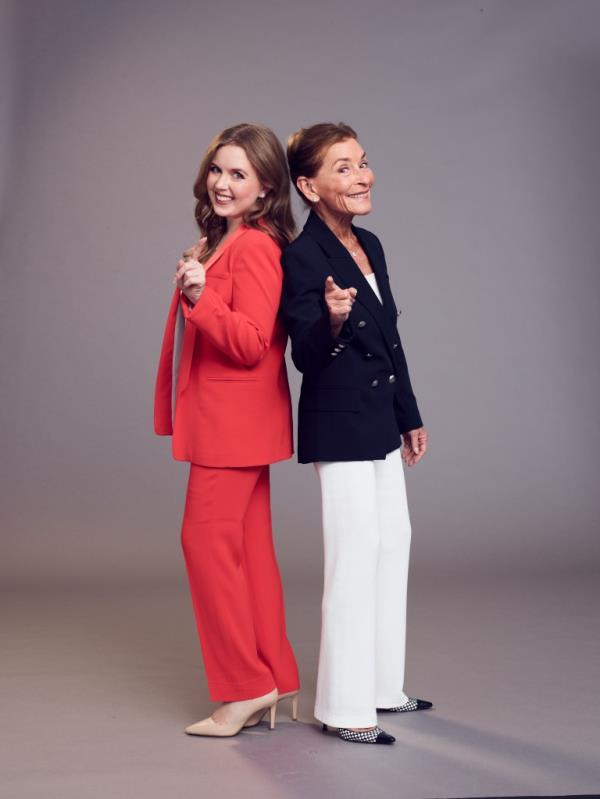 Sarah Rose wearing a red pantsuit holding up her pointer finger standing back to back with her grandmother, Judge Judy Sheindlin who is wearing a black blazer and white dress pants holding the same pose. 