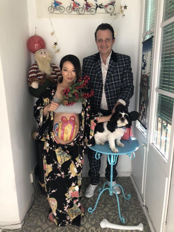 A photo of Makiyo and Edward Davidson with Panda, with a pregnant makiyo with her belly exposed and a painting of a  bright yellow gift box with a red ribbon on her stomach