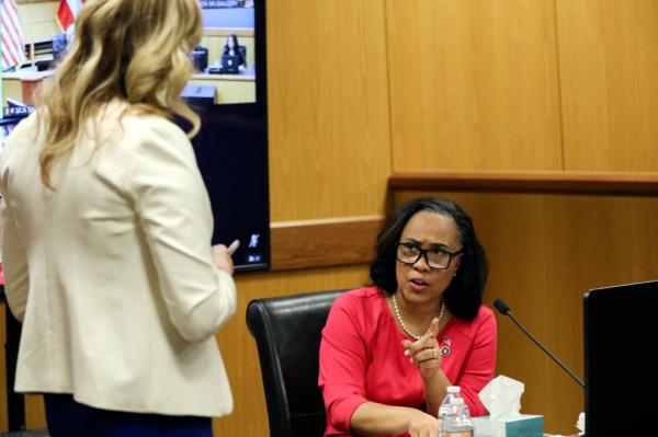 A photo of Fani Willis in a red dress and Ashleigh Merchant in a cream-colored suit talking in the courtroom.