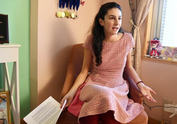 Kristina Raevsky sitting in chair in bedroom wearing pink dress, holding papers, talking emphatically while looking off camera; debate medals seen behind her