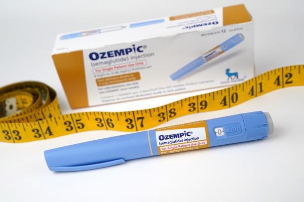 A pic of box of Ozempic in the background with a yellow/orange tape measure in front of it and then an Ozempic pen in front of that.