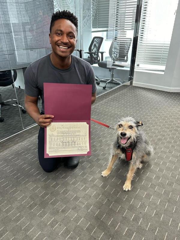 Councilman Chi Ossé kneels next to Luna, a small Schnauzer, and holds a certificate. 