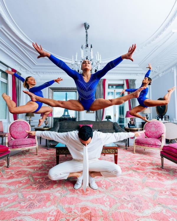 A group of people in a room with a chandelier, featuring a woman in a blue leotard jumping mid-air at Plaza Athenee hotel Paris Olympic team event, photographed by Mathieu Forget
