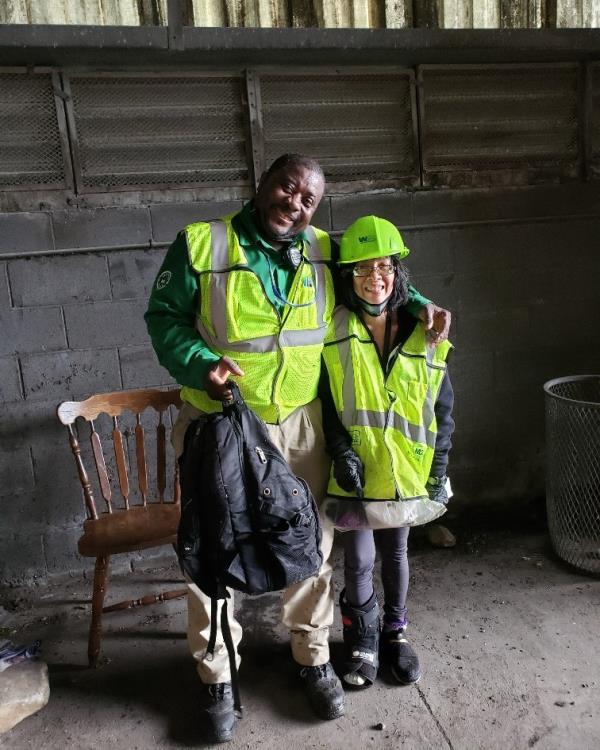 A sanitation worker holds a black backpack and puts his arm around a woman. Both smile and wear bright yellow vests.  