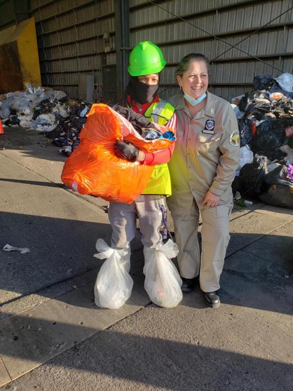 A person holds an orange garbage bag filled with stuff, while wearing a mask and green hard helmet. A woman smiles next to her. trash in the background. 
