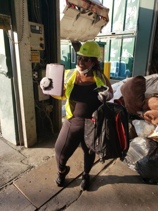 A woman wears a hard helmet and yellow vest and smiles while holding a laptop in one hand and a backpack in the other. 