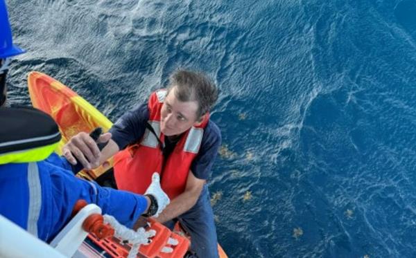 A person reaches down to grab a man climbing up a ladder on the side of large ship as a kayak in the ocean water can be seen below.