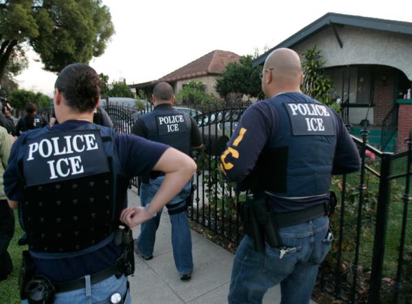 Three ICE police officers walk along a residential street in dark blue work uniforms that read 