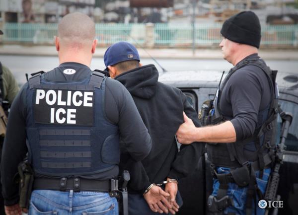 Two ICE police officers lead. a handcuffed man into a car