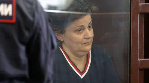 BBC Anna sitting behind the glass screen of the court cage with a wooden f<em></em>rame. She is on the right side of the shot, looking towards the right with a serious ex<em></em>pression on her face. She has short dark hair, wearing a navy v neck jumper with red trim. On the left-hand is the back of a security guard with Russian writing on his navy uniform.