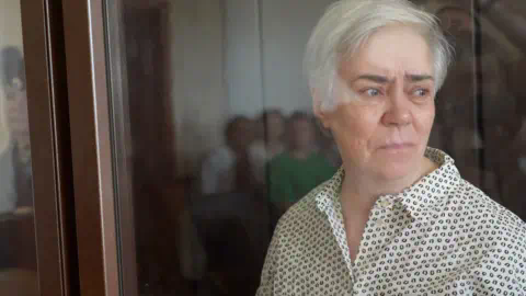 Nadezhda Buyanova, who is also pictured behind the glass of a court dock, looks to the right with a serious ex<em></em>pression on her face. She has short grey hair and dark eyebrows, wearing a white shirt with a black dotted pattern.